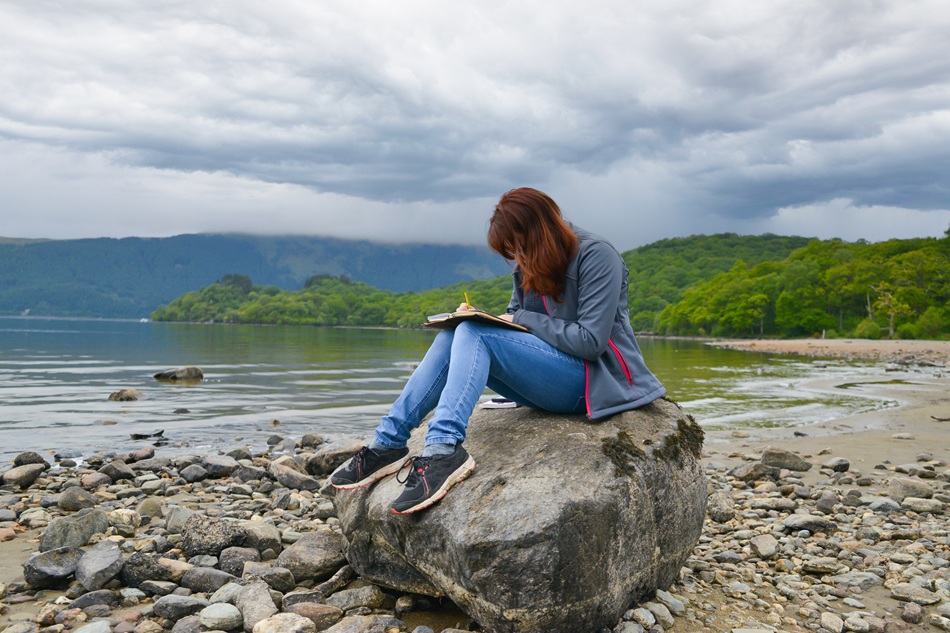 woman writing in a journal sat on a beach next to a loch